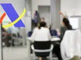 Logo de Hacienda en primer plano. Al fondo, personas atendiendo trámites, quizás esperando cuando devuelve Hacienda.