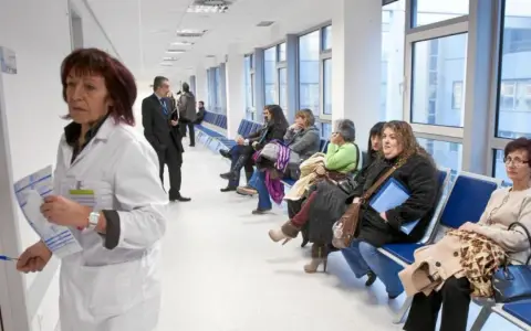 Doctora con bata blanca revisa papeles, mientras pacientes esperan en pasillo. Quizás pensando en cómo pedir excedencia estando de baja.