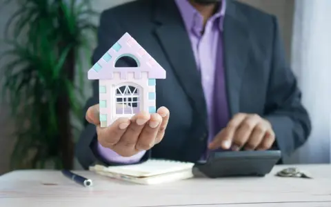 Hombre con traje calcula cuánto puede subir el alquiler, sosteniendo una casa de juguete.