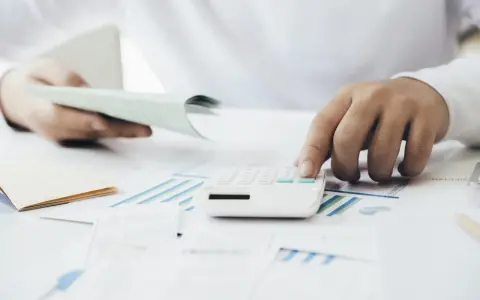 Manos calculando con calculadora blanca sobre gráficos y papeles. Una mano sostiene un talonario, la otra opera la calculadora. Proceso de factura simplificada.