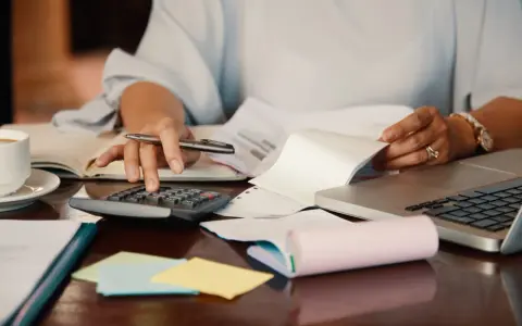 Persona revisa documentos y usa calculadora para el modelo 184. Hay café, portátil y notas adhesivas.