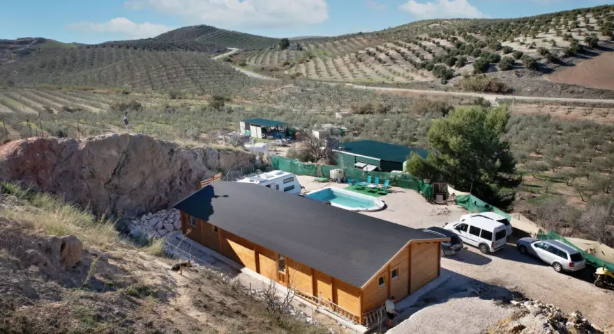 Casa de madera con piscina y caravana en terreno rústico, rodeada de olivos y colinas. Ideal para quienes buscan un refugio natural.