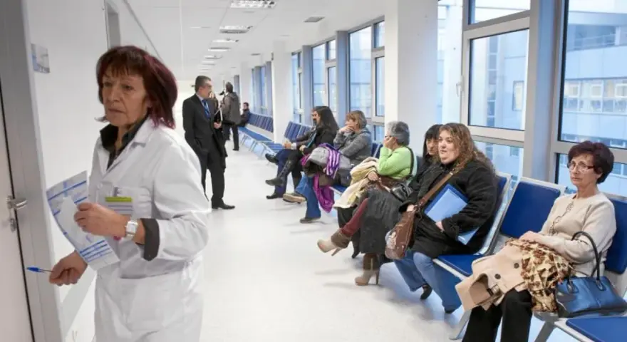 Doctora con bata blanca revisa papeles, mientras pacientes esperan en pasillo. Quizás pensando en cómo pedir excedencia estando de baja.
