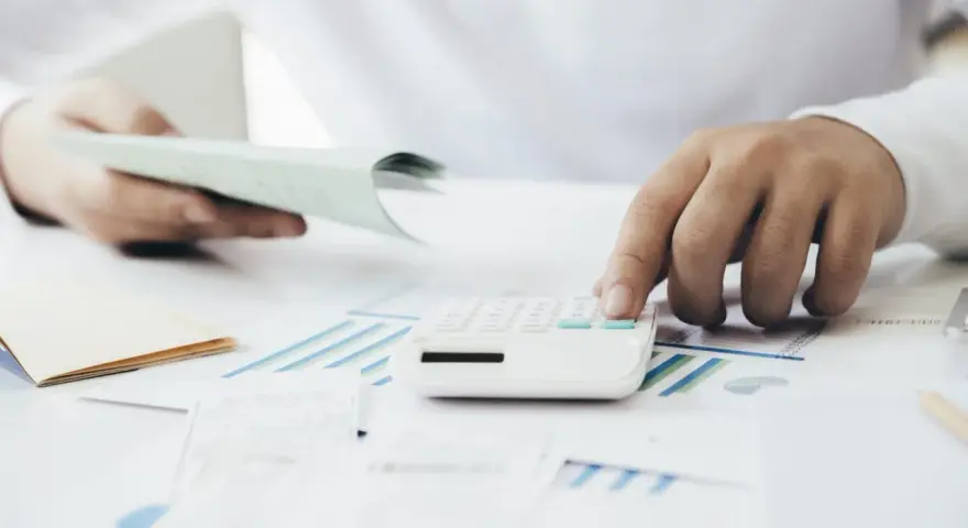 Manos calculando con calculadora blanca sobre gráficos y papeles. Una mano sostiene un talonario, la otra opera la calculadora. Proceso de factura simplificada.