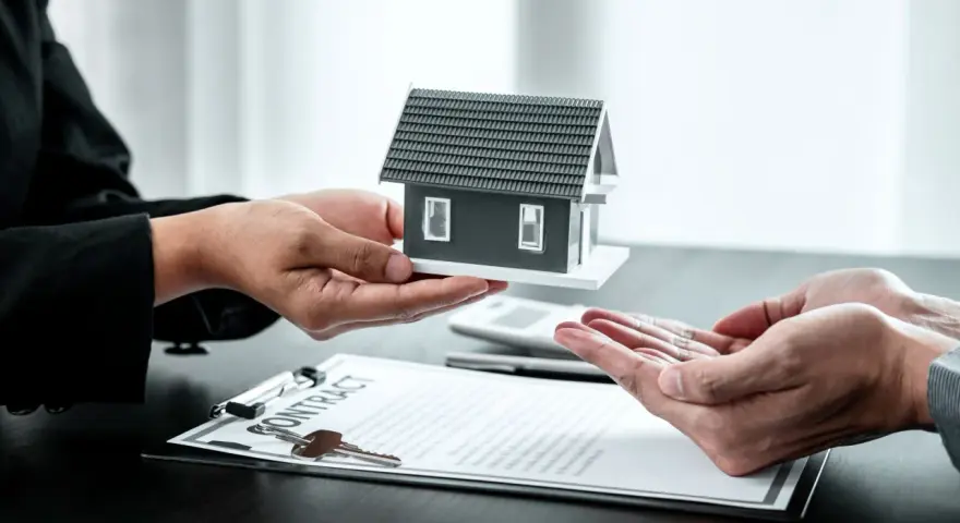 Manos entregan maqueta de casa sobre contrato y llaves. Posible exención IRPF venta vivienda heredada.