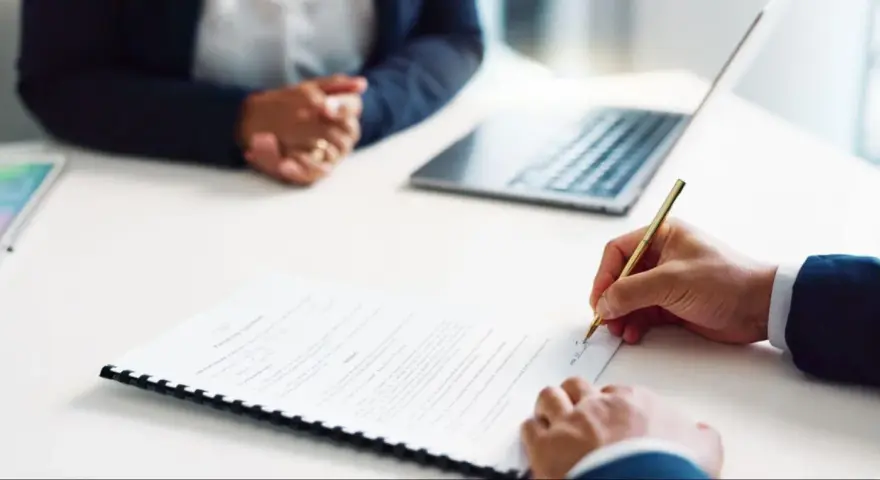 Persona firma un contrato de alquiler de habitación. A su lado, otra persona espera, con un portátil y una tablet sobre la mesa.