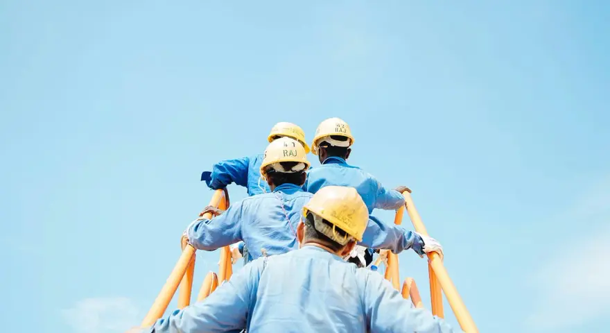 Trabajadores con cascos amarillos suben una escalera naranja contra un cielo azul claro. Podría ser una escena de cesión ilegal de trabajadores.