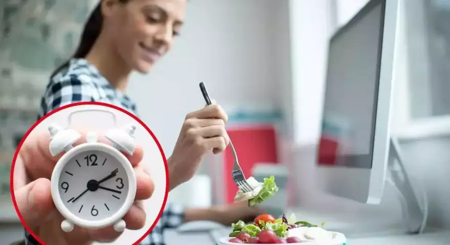 Mujer comiendo ensalada en su escritorio, con un reloj que marca el tiempo para comer en el trabajo.