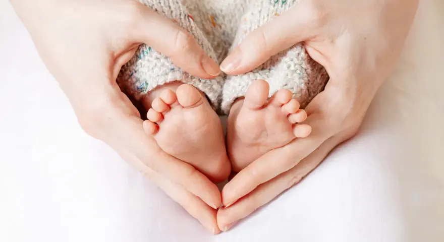 Manos formando un corazón alrededor de los pies de un bebé, evocando la protección y el complemento de maternidad con retroactividad.