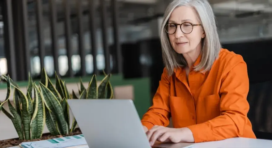 Mujer mayor con cabello plateado y gafas trabajando en su laptop, un ejemplo de jubilación activa.