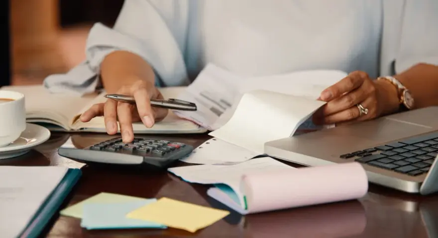 Persona revisa documentos y usa calculadora para el modelo 184. Hay café, portátil y notas adhesivas.