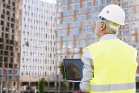 Ingeniero con casco y chaleco reflectante revisa un portátil frente a edificios en construcción. A pesar de un posible **ite desfavorable**, su mirada es decidida.