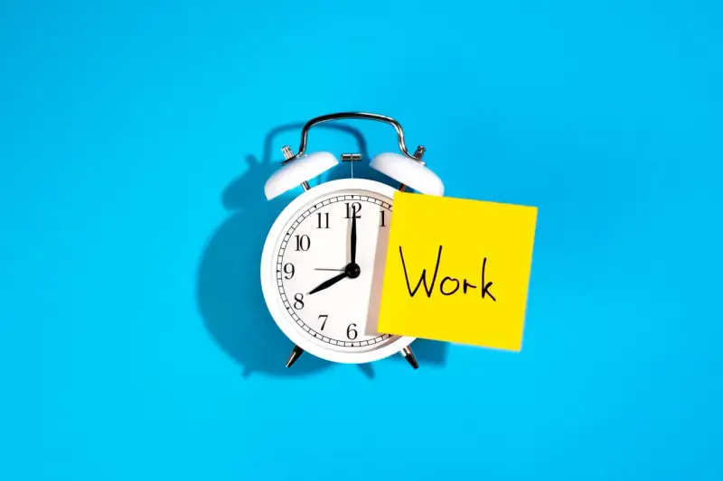 Reloj despertador blanco con nota amarilla que dice "Work", recordando la importancia de la jornada laboral.