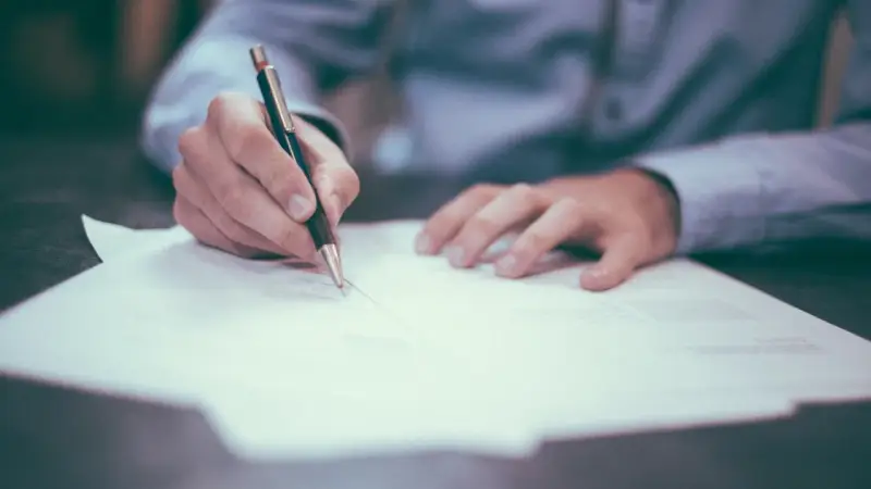 Mano firmando un contrato de reserva de vivienda. El documento se extiende sobre una mesa, con un bolígrafo en acción.